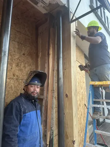 Workers beside a boarded storefront opening and vertical support posts during structural repair.