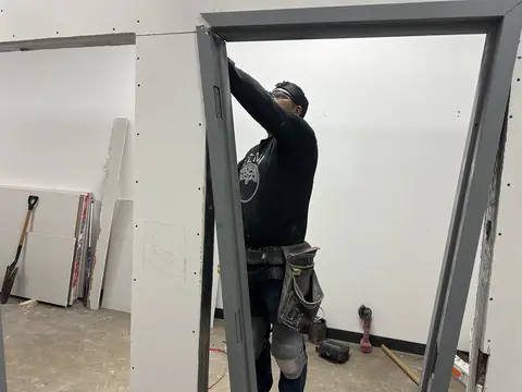 A worker fitting a metal door frame into an unfinished drywall opening.