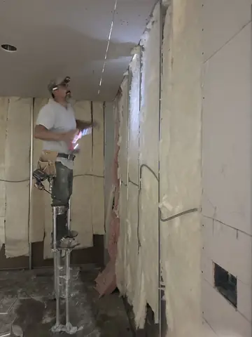 A worker on stilts working on ceiling drywall beside exposed wall insulation.