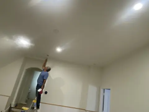 Ceiling finishing from a stepladder above a room with wall paneling and trim.